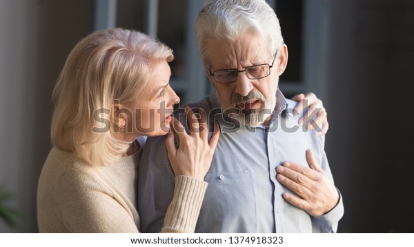 Grey haired man touching chest, having heart attack,\
feeling pain, suffering from heartache disease at home, mature\
woman supporting, embracing him, middle aged family, horizontal\
banner, close up