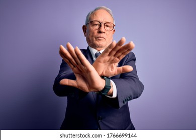 Grey haired senior business man wearing glasses and elegant suit and tie over purple background Rejection expression crossing arms and palms doing negative sign, angry face