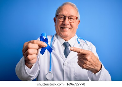 Grey Haired Senior Doctor Man Holding Colon Cancer Awareness Blue Ribbon Over Blue Background Very Happy Pointing With Hand And Finger