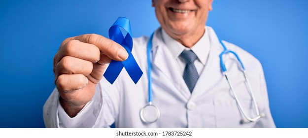 Grey Haired Senior Doctor Man Holding Colon Cancer Awareness Blue Ribbon Over Blue Background With A Happy Face Standing And Smiling With A Confident Smile Showing Teeth