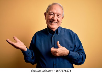 Grey haired senior man wearing casual blue shirt standing over yellow background Showing palm hand and doing ok gesture with thumbs up, smiling happy and cheerful