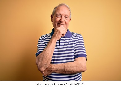 Grey haired senior man wearing casual navy striped t-shirt standing over yellow background looking confident at the camera with smile with crossed arms and hand raised on chin. Thinking positive.