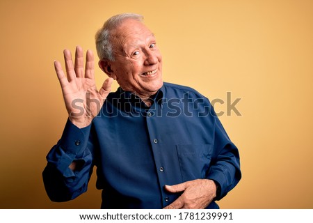 Grey haired senior man wearing casual blue shirt standing over yellow background Waiving saying hello happy and smiling, friendly welcome gesture
