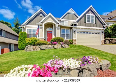 Grey House Exterior With Entrance Porch And Red Door. Beautiful Front Yard Landscape With Vivid Flower And Stones