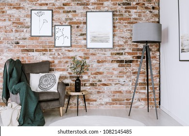 Grey lamp and armchair with white pillow set on a brick wall with posters in living room interior