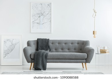 Grey Lounge With Dark Blanket Standing In White Living Room Interior With Two Map Posters, Gold Bulb Lamp And Books On A Small Table