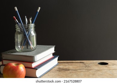 Grey, Red And Navy Blue Books Stacked On An Old Desk With A Red Apple And A Jar Of Pens With Copy Space On Black Background