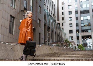 Grey Senior Woman In Leather Jacket Holding Bag While Going Up Stairs Outdoors