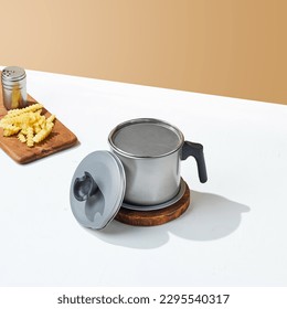grey, steel-based cooking oil strainer, with fries surround it on a white and cream background.