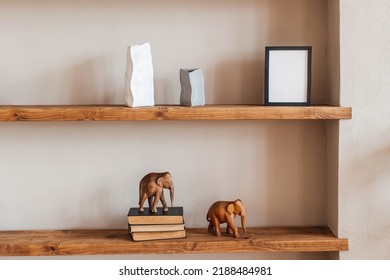 Grey And White Colored Vases, Wooden Figures Of Elephants, Blank Black Photoframe And Stack Of Books On Wooden Shelf Against  Grey Wall. Home Decor.