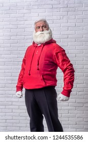 A grey-haired bearded man in a red jacket in the image of Santa Claus is standing against a white wall and posing emotionally.