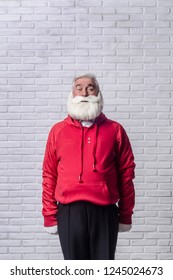 A grey-haired bearded man in a red jacket in the image of Santa Claus is standing against a white wall and posing emotionally.