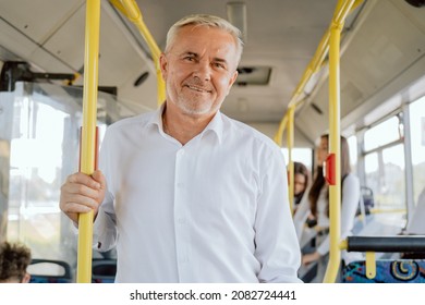 Grey-haired Elderly Man Dressed In Smart White Shirt Stands In Middle Of Bus, He Takes Public Transport To Work, Guy Is Holding Onto Railing Next To Door Opening Button, He Smiles At The Camera