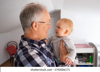 Grey-haired Grandpa In Eyeglasses Holding Cute Baby Girl. Grandfather And Grandchild Looking At Each Other. Help From Grandparents And Staying At Home Concept