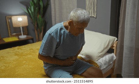 A grey-haired hispanic man in pajamas sitting on bed holding his stomach in a cozy bedroom, displaying signs of discomfort, with a nightstand lamp illuminating the room. - Powered by Shutterstock