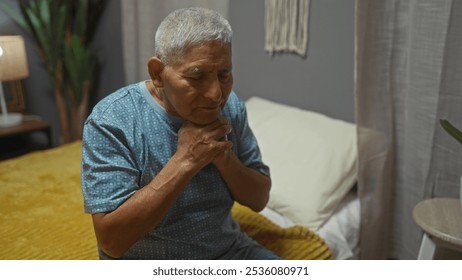 A grey-haired hispanic man sits in a bedroom looking thoughtful, wearing a blue shirt, with a yellow bedspread and home decor visible in the indoor setting. - Powered by Shutterstock