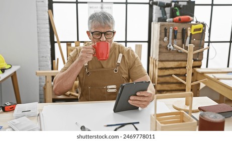 A grey-haired man enjoys a coffee break in a woodworking workshop, browsing on a tablet amidst handcrafted woodwork. - Powered by Shutterstock