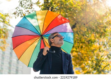 Grey-haired middle-aged man in formal clothes walking around city park holding rainbow umbrella looking up. Horizontal outdoor shot. High quality photo