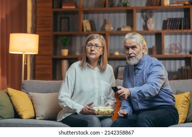 Grey-haired Older Couple At Home Choose A Television Show To Watch And Switch Channels With A TV Remote Control. Elderly Mature Family Husband And Wife Cannot Choose A Show. Watching News Or Film
