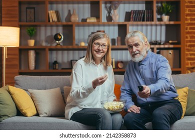 Grey-haired Older Couple At Home Choose A Television Show To Watch And Switch Channels With A TV Remote Control. Elderly Mature Family Husband And Wife Cannot Choose A Show. Watching News Or Film