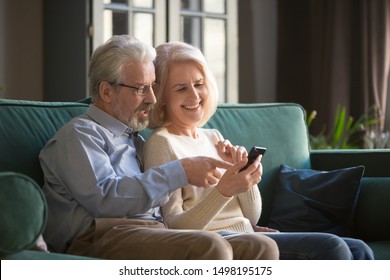 Grey-haired pensioner in eyeglasses helping his aged wife with new smartphone app, hugging. Happy mature family spouse using cell phone together at home, surfing internet, sitting on couch. - Powered by Shutterstock