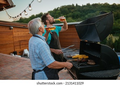 Grey-haired pensioner spending time with son while drinking beer and barbecuing. Both of them wearing apron