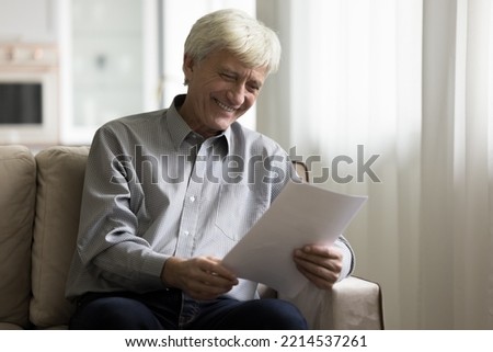 Grey-haired senior man smiling while reading paper letter, get good news in bank notification seated on sofa at home. Pleasant notice, finances growth, good terms and conditions in commercial offer