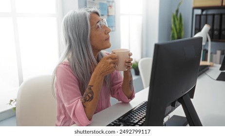 Grey-haired woman sipping coffee in an office, seated at a desk with tattoos visible, reflecting the modern and diverse workplace, enclosed by computers and office supplies. - Powered by Shutterstock