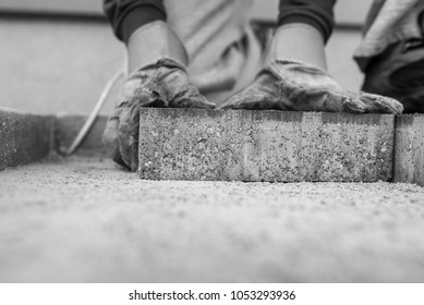 Greyscale image of the gloved hands of a manuel worker laying outdoor paving slabs on a prepared base.