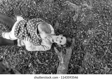 Greyscale image of smiling mother playing with her baby girl on the grass amongst autumn leaves, top view.