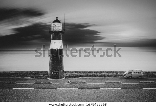 A greyscale shot of a\
lighthouse on the road with a van on the side and the sea on the\
background
