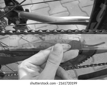 Greyscaled image - Close view on manual checking of bike chain wear and tear degree by measuring the length of segments with a transparent plastic ruler.