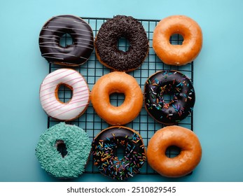 A grid of assorted donuts is displayed on a black cooling rack against a bright turquoise background. The donuts include glazed, chocolate, sprinkle-topped, and coconut-covered varieties. - Powered by Shutterstock