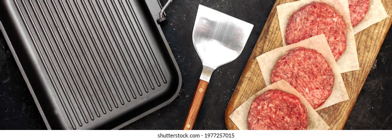 Griddle Grill Pap And Ground Beef Meat Patties For Grilling On Black Background, Overhead View. Raw Steak Burgers Cutlets On Grill Pan.  Burgers For Frying, Grill Tools And Cutting Board, Top View.