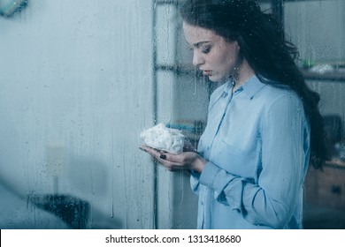 Grieving Mother Holding Baby Shoes Through Window With Raindrops And Copy Space