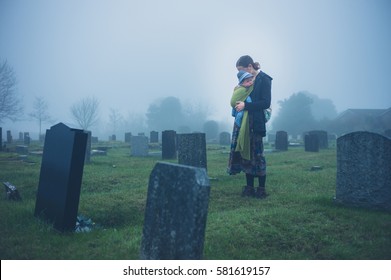 A Grieving Young Mother Is Standing With Her Baby By A Tombstone In A Graveyard