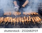 A grill filled with skewers of meat cooking over open flames. The meat browns, while someone tends to the grill wearing gloves. The scene shows a traditional barbecue in progress.Greek Souvlaki