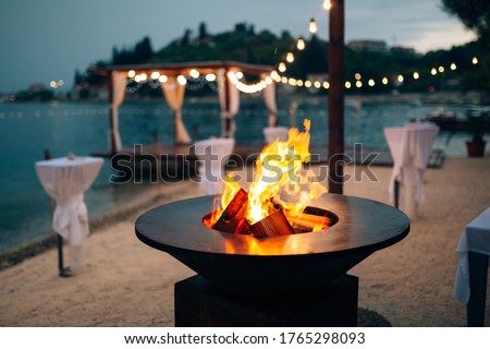 Grill with flames inside. Round table-cooking surface. On the beach, in the background of the gazebo by the water with garlands, in the twilight light.