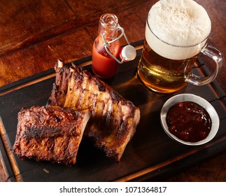 Grilled or barbecued spicy ribs with a cold beer with frothy head served on a tray with chutney and tomato ketchup for a tasty finger snack pub lunch