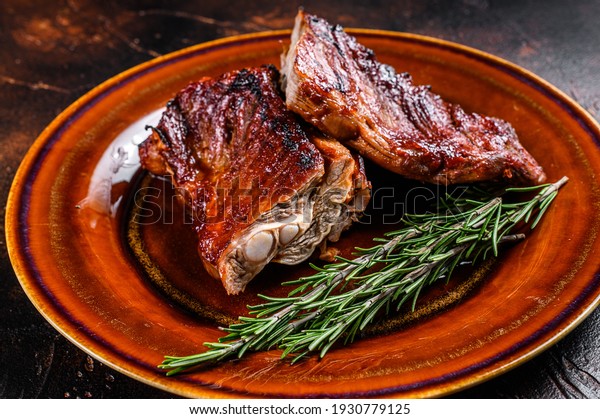 Grilled BBQ veal calf short spare rib meat on\
plate. Dark background. Top\
view.