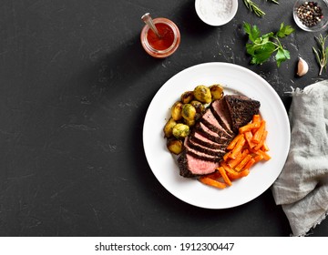 Grilled beef steak with brussels sprouts and sweet potatoes on plate over black stone background with free text space. Top view, flat lay