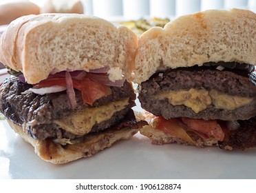 A Grilled Burger Stuffed With Cheese On A Hoagie Bun And Topped With Onion , Tomato And Pickle Is Cut Open Showing The Cheese Melting Out Of It