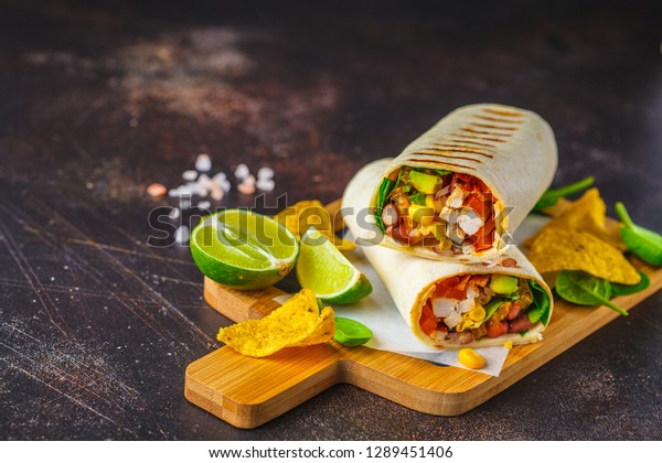 Grilled burritos wraps with chicken, beans, corn,\
tomatoes and avocado on a wooden board, dark background. Meat\
burrito, mexican food.