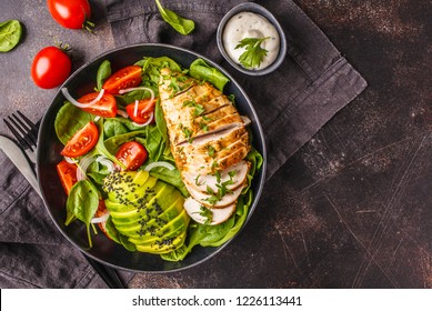 Grilled chicken breast and avocado salad with spinach, tomatoes and Caesar dressing in black plate, dark background.