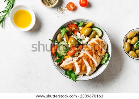 Grilled chicken fillet and fresh vegetable greek salad on white, top view, copy space. Healthy keto, ketogenic meal - roasted chicken breast meat, olives, feta cheese, vegetables and greens.
