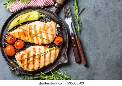 Grilled chicken fillets, chili pepper and tomatoes cherry on grill iron pan, fresh herbs - rosemary and parsley, rustic cutlery on gray slate background. top view. copy space