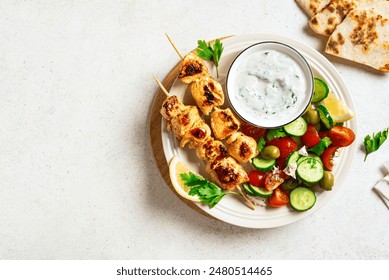 Grilled chicken kebab with fresh vegetable salad on white background, top view, copy space. Mediterranean lunch with roasted skewers, tzadziki sauce and veggies, healthy food.