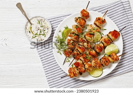 grilled chicken onion skewers with thyme and lime wedges on white plate on white wooden table with tzatziki sauce, horizontal view from above, flat lay, free space
