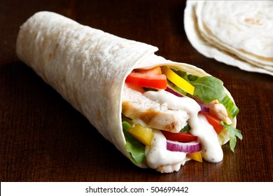 Grilled Chicken And Salad Tortilla Wrap With White Sauce Isolated On Dark Background. Next To Pile Of Tortillas.