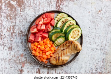 Grilled chicken zucchini carrot tomato vegetables salad in plate on white wood background top view, healthy food concept.  